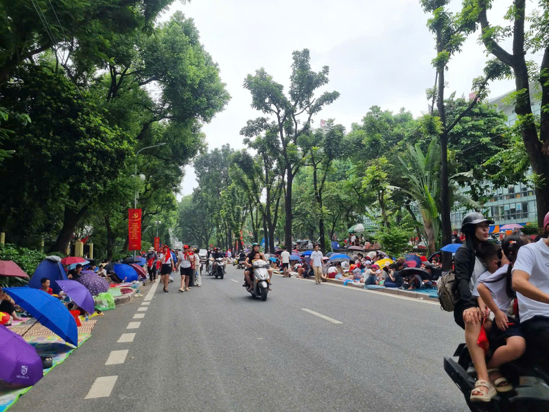 Trên đường Kim Mã, từng tốp người ngồi ken đặc vỉa hè, mang theo cờ hoa và ô che nắng, kiên nhẫn chờ đoàn diễu binh sự kiện A80 đi qua.