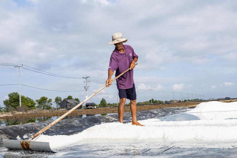 Dưới nắng gió Lý Nhơn, diêm dân mạnh mẽ đẩy từng lớp nước mặn, gom lại thành hạt muối trắng sau nhiều giờ phơi mình giữa ruộng đồng. Công việc lặp đi lặp lại mỗi ngày, nặng nhọc nhưng chưa từng ngưng nghỉ, như cách diêm dân gìn giữ nghề truyền thống qua năm tháng. Ảnh: Hà Trang